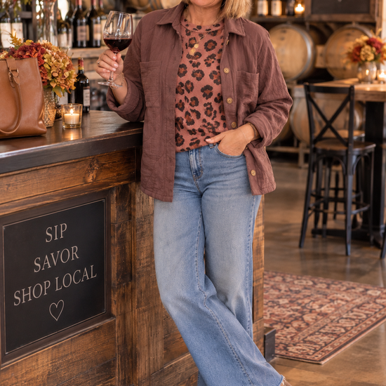 Woman styling a mauve button-front jacket over a leopard-print top with wide-leg jeans, ankle boots, and a felt hat at a winery