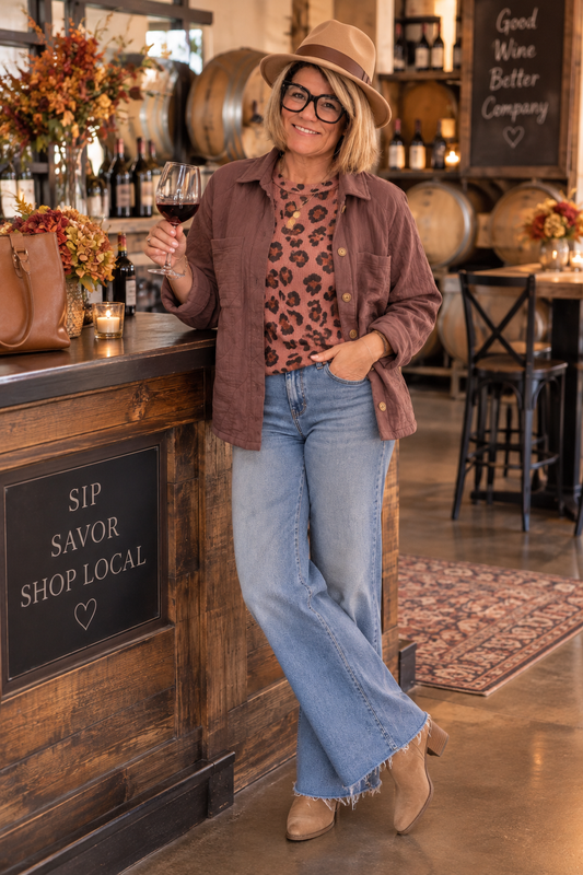 Woman styling a mauve button-front jacket over a leopard-print top with wide-leg jeans, ankle boots, and a felt hat at a winery