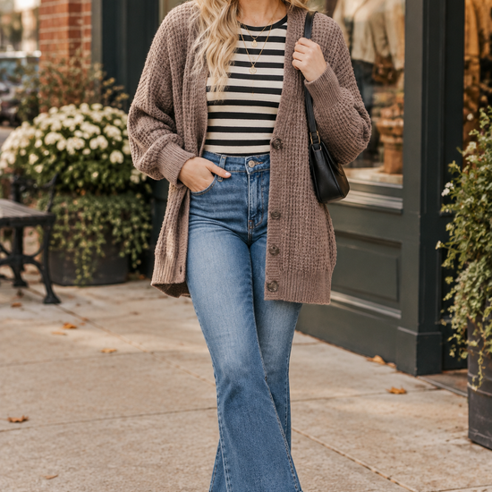 Woman wearing a taupe chunky knit cardigan over a striped long-sleeve shirt with cropped flare jeans and white sneakers